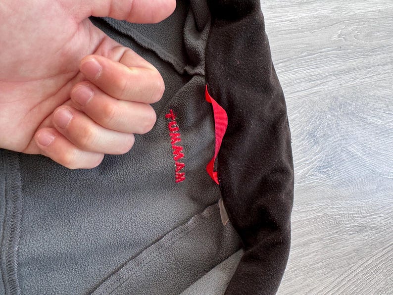 May include: Close-up of a gray and black fleece jacket. The jacket has a red pull tab and the word "TOM MAX" embroidered in red on the gray fabric. The jacket is made of a soft, textured material.