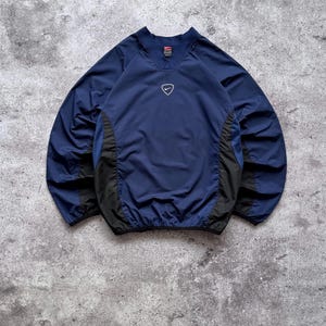 May include: A navy blue long-sleeve pullover windbreaker featuring a V-neck collar, black side panels, and a small white Nike swoosh logo centered on the chest. The garment has elastic cuffs and a hem, with a Nike brand label inside the collar.