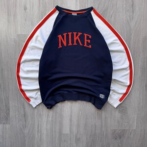 May include: Navy blue sweatshirt with white sleeves and red accents. The word "NIKE" is displayed in large, red, block letters across the chest. A small Nike logo is visible on the lower right.