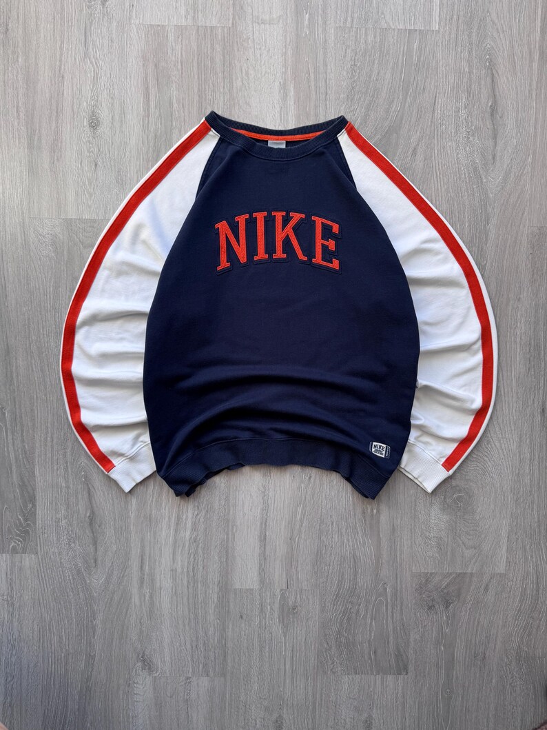 May include: Navy blue sweatshirt with white sleeves and red accents. The word "NIKE" is displayed in large, red, block letters across the chest. A small Nike logo is visible on the lower right.