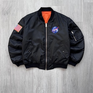 Puede incluir: Chaqueta bomber negra con cierre de cremallera, con un parche con el logotipo de la NASA y un parche con la bandera estadounidense en la manga. La chaqueta tiene cuello, puños y dobladillo de canalé negro, con forro naranja.