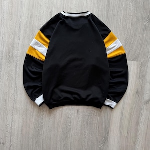 Puede incluir: Sudadera negra con rayas blancas y amarillas en las mangas. El cuello y los pu&ntilde;os tienen ribetes blancos. La prenda est&aacute; tendida sobre una superficie de madera gris claro.