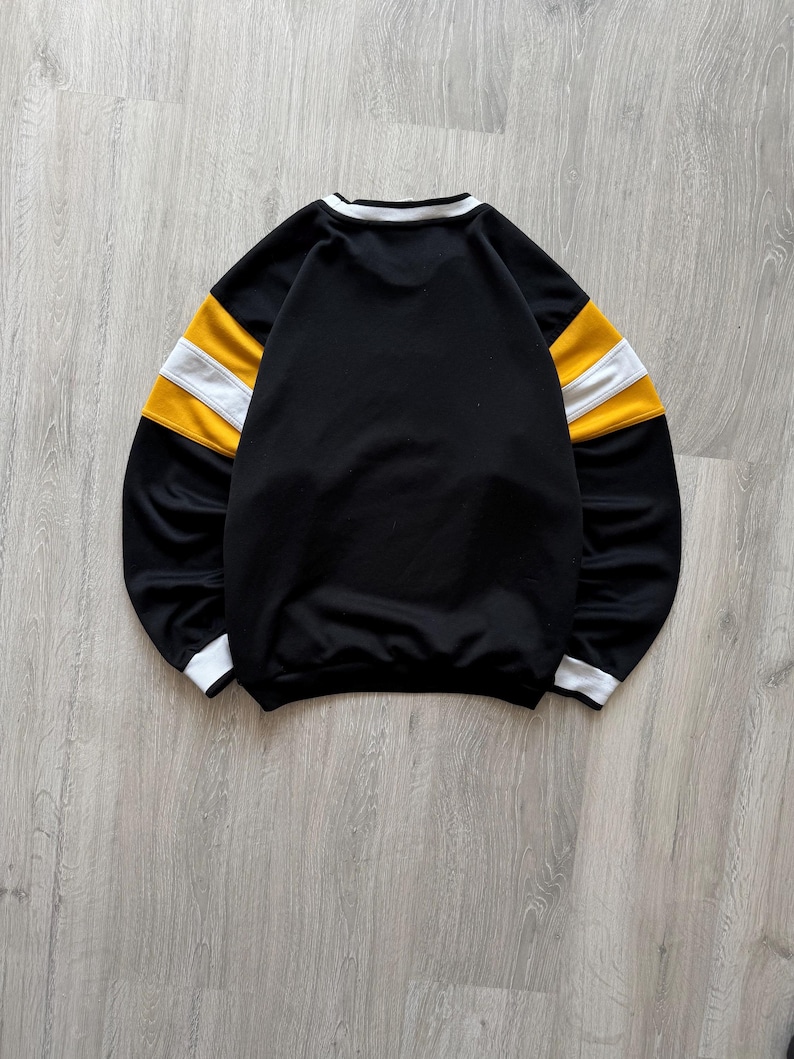 Puede incluir: Sudadera negra con rayas blancas y amarillas en las mangas. El cuello y los pu&ntilde;os tienen ribetes blancos. La prenda est&aacute; tendida sobre una superficie de madera gris claro.