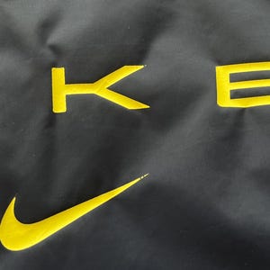 May include: Black fabric with yellow embroidered Nike swoosh and the letters "KE" in a bold, sans-serif font. The fabric has a slight sheen, and the yellow embroidery provides a striking contrast against the dark background.