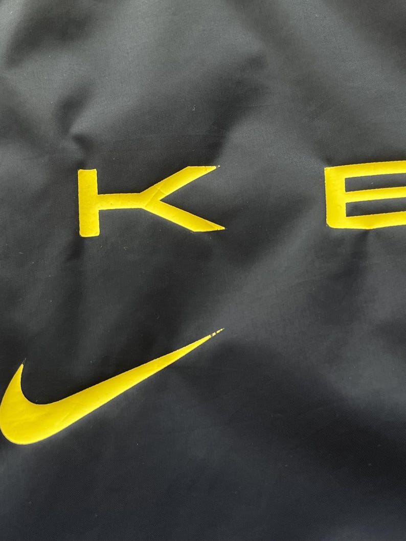 May include: Black fabric with yellow embroidered Nike swoosh and the letters "KE" in a bold, sans-serif font. The fabric has a slight sheen, and the yellow embroidery provides a striking contrast against the dark background.