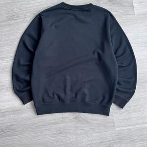 Peut inclure: Un sweat-shirt noir uni avec un col rond et des poignets et un ourlet c&ocirc;tel&eacute;s. Le v&ecirc;tement est fait d'une mati&egrave;re douce et est pr&eacute;sent&eacute; sur une surface en bois gris clair. Un v&ecirc;tement simple et polyvalent.