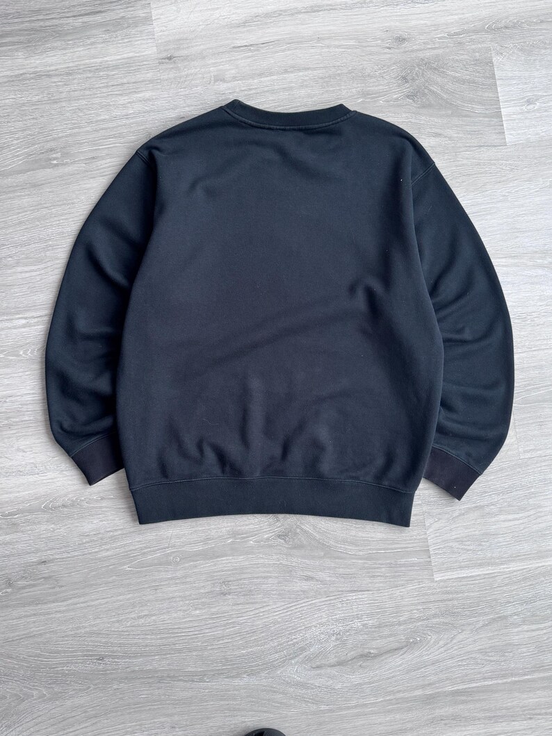 Peut inclure: Un sweat-shirt noir uni avec un col rond et des poignets et un ourlet c&ocirc;tel&eacute;s. Le v&ecirc;tement est fait d'une mati&egrave;re douce et est pr&eacute;sent&eacute; sur une surface en bois gris clair. Un v&ecirc;tement simple et polyvalent.