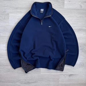 May include: A navy blue fleece pullover with a quarter-zip closure and a small white Nike logo on the chest. The pullover has a high collar and black accents on the zipper and side pockets.