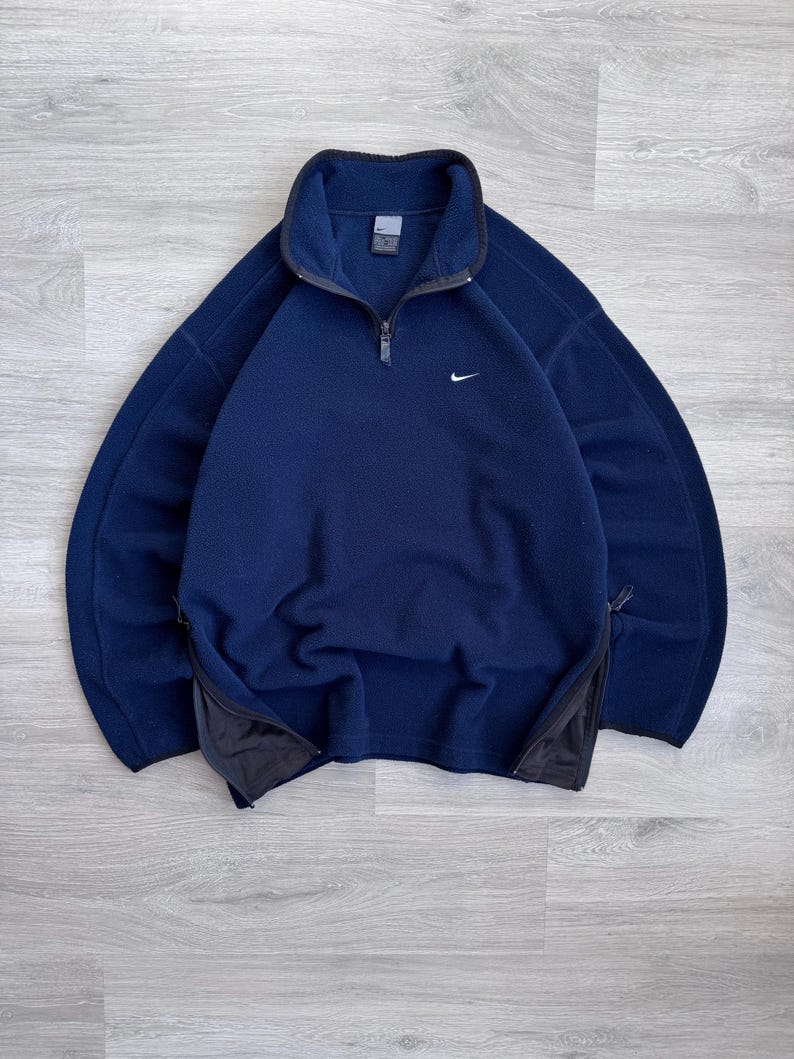 May include: A navy blue fleece pullover with a quarter-zip closure and a small white Nike logo on the chest. The pullover has a high collar and black accents on the zipper and side pockets.