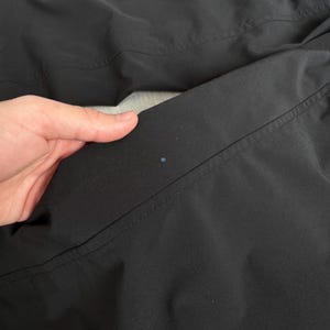 Puede incluir: Primer plano de tela negra con costuras visibles y un peque&ntilde;o punto azul. La tela parece ser parte de una prenda, posiblemente pantalones o una chaqueta, con un acabado liso y mate.