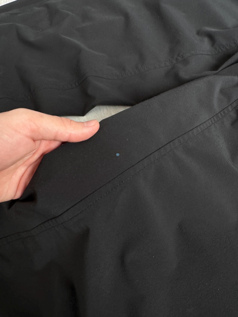 Puede incluir: Primer plano de tela negra con costuras visibles y un peque&ntilde;o punto azul. La tela parece ser parte de una prenda, posiblemente pantalones o una chaqueta, con un acabado liso y mate.