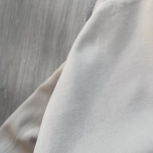 May include: A close-up shot of a beige sweatshirt. The fabric appears soft and smooth, with visible stitching along the neckline and sleeve. The neutral color suggests a versatile garment suitable for various outfits.