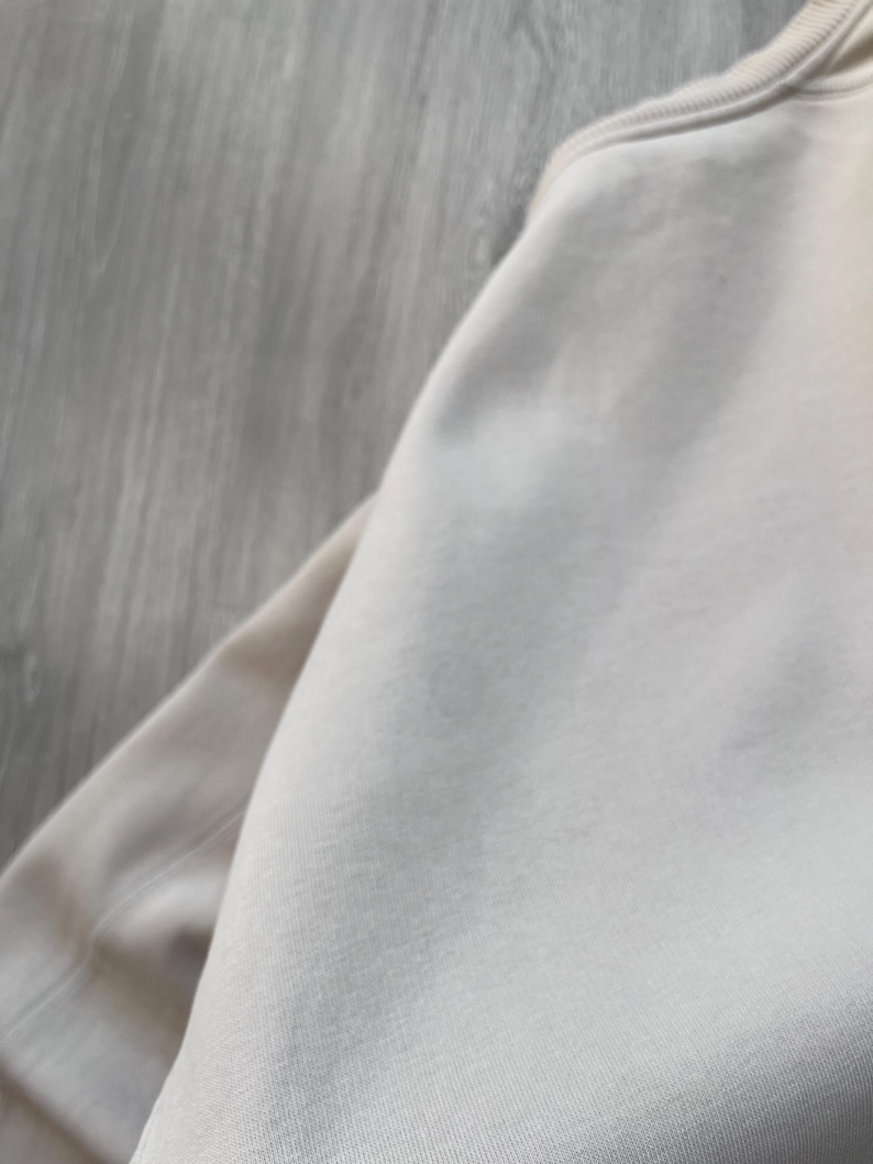 May include: A close-up shot of a beige sweatshirt. The fabric appears soft and smooth, with visible stitching along the neckline and sleeve. The neutral color suggests a versatile garment suitable for various outfits.