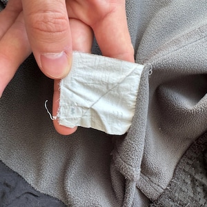 May include: Close-up of a gray fleece jacket with a white tag. The tag is held by a hand and is attached to the jacket's collar. The jacket has a black trim and a textured pattern on the lower right side.
