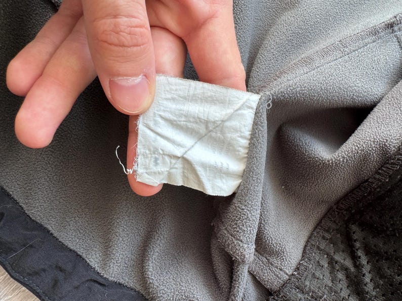 May include: Close-up of a gray fleece jacket with a white tag. The tag is held by a hand and is attached to the jacket's collar. The jacket has a black trim and a textured pattern on the lower right side.