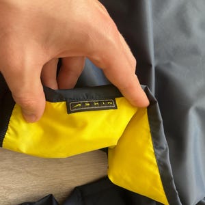 May include: Navy blue jacket with a yellow interior. The jacket has a black ribbed collar and cuffs. A small rectangular tag with the brand name "NIKE" is visible.