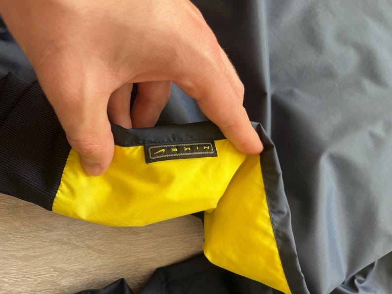 May include: Navy blue jacket with a yellow interior. The jacket has a black ribbed collar and cuffs. A small rectangular tag with the brand name "NIKE" is visible.