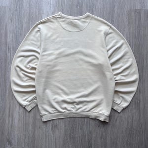 Puede incluir: Una sudadera de color crema con cuello redondo y mangas largas. La parte trasera de la sudadera es visible, mostrando el escote y la tela suave y texturizada. Los pu&ntilde;os y el dobladillo son acanalados.