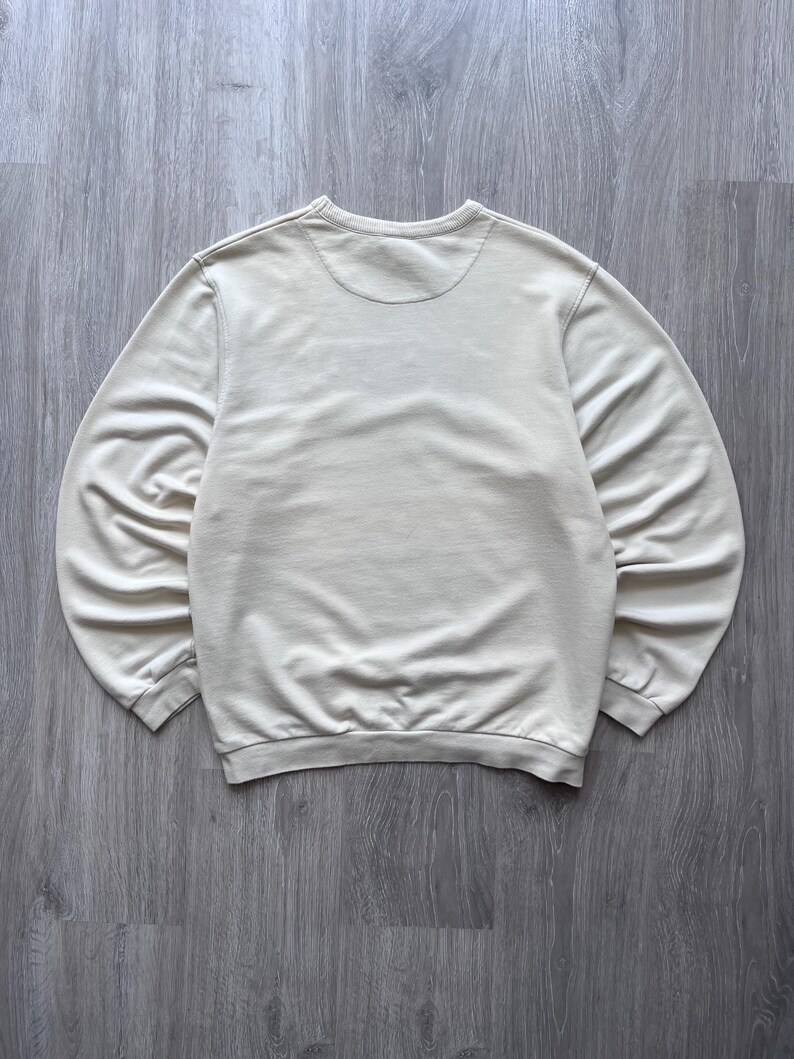 Puede incluir: Una sudadera de color crema con cuello redondo y mangas largas. La parte trasera de la sudadera es visible, mostrando el escote y la tela suave y texturizada. Los pu&ntilde;os y el dobladillo son acanalados.