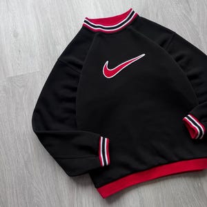May include: Black sweatshirt with red and white striped detailing on the collar, cuffs, and hem. A red and white Nike swoosh logo is on the chest. The sweatshirt has a classic athletic design.