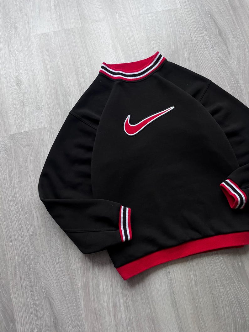 May include: Black sweatshirt with red and white striped detailing on the collar, cuffs, and hem. A red and white Nike swoosh logo is on the chest. The sweatshirt has a classic athletic design.