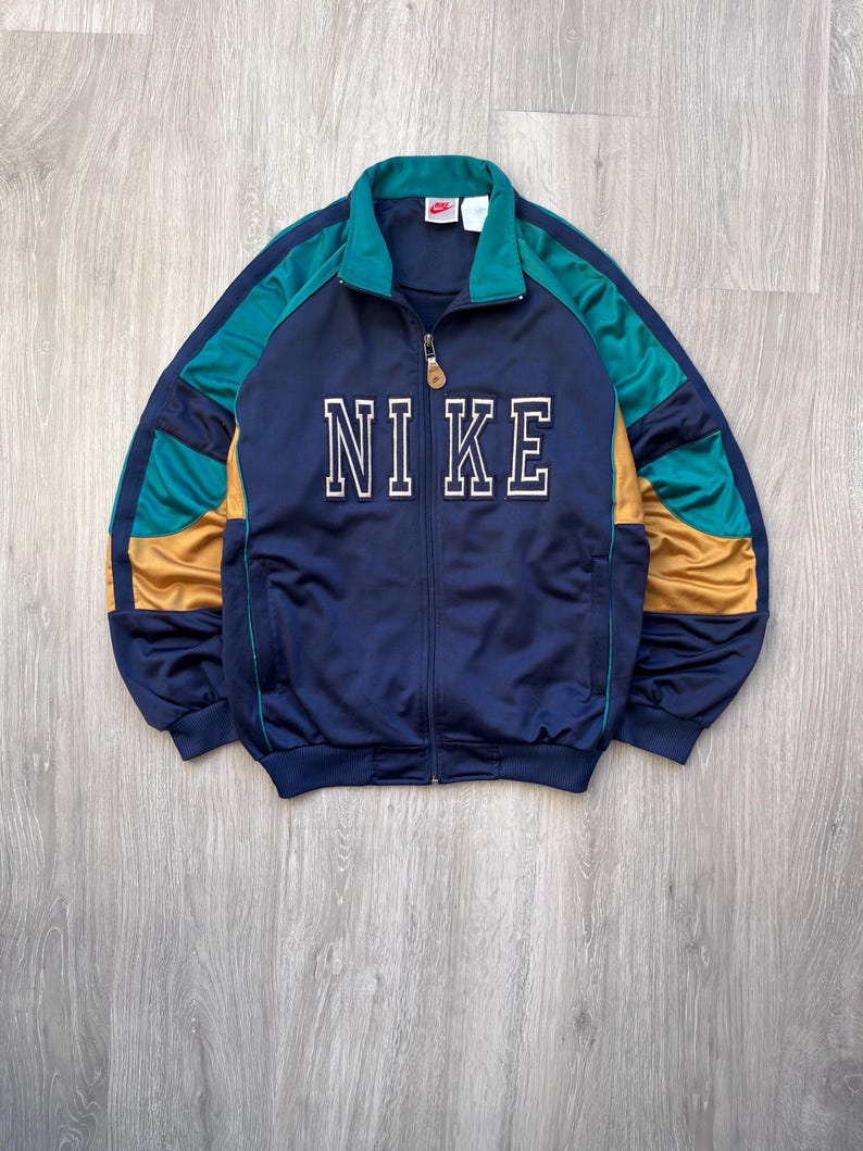 May include: A navy blue Nike track jacket with teal and gold accents. The jacket features a full zipper and the Nike logo in white block letters across the chest. The jacket has a ribbed collar and cuffs.