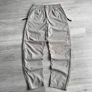 May include: A pair of light grey cargo trousers with black zipper accents and a black Nike swoosh logo on the right leg.