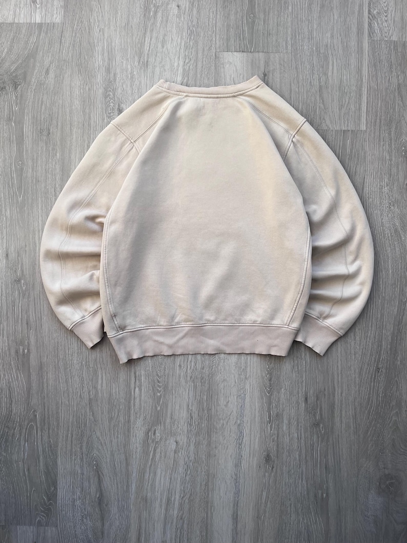 May include: A beige crewneck sweatshirt, laid flat on a wooden surface. The sweatshirt has a ribbed hem and cuffs, with a simple, classic design. The fabric appears soft and slightly worn.