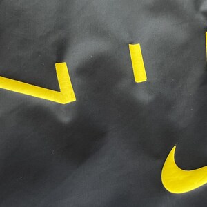 May include: A dark gray fabric with yellow cut-out shapes, including a curved checkmark and several straight lines. The shapes are arranged in a scattered pattern across the surface.