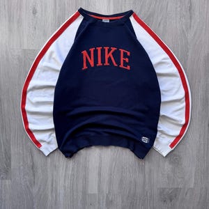 May include: Navy blue sweatshirt with the word "NIKE" in red block letters across the chest. The sleeves are white with a red stripe down the side. A small tag is visible on the bottom hem.