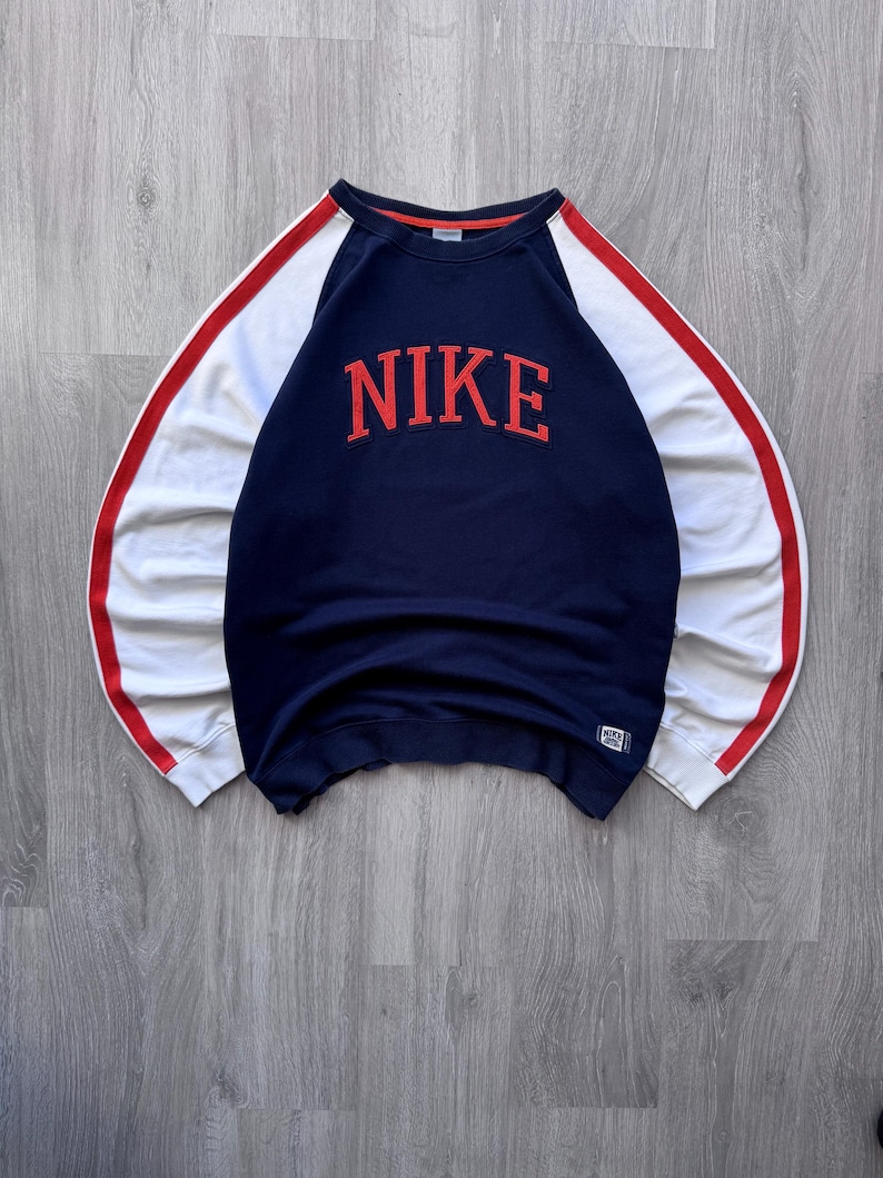 May include: Navy blue sweatshirt with the word "NIKE" in red block letters across the chest. The sleeves are white with a red stripe down the side. A small tag is visible on the bottom hem.