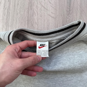 May include: A gray Nike sweatshirt with a black Nike swoosh logo and a black and white striped collar. The tag reads "Nike" and includes size information. The sweatshirt is made in Greece.