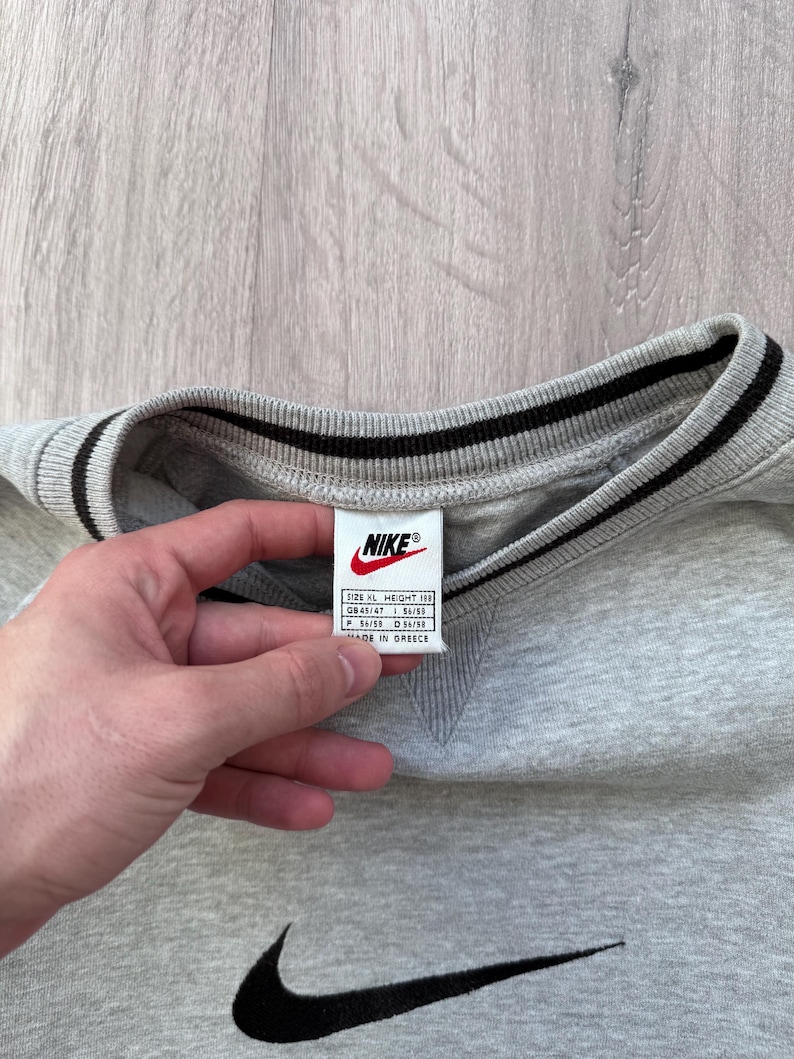 May include: A gray Nike sweatshirt with a black Nike swoosh logo and a black and white striped collar. The tag reads "Nike" and includes size information. The sweatshirt is made in Greece.