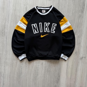 Puede incluir: Sudadera negra Nike con ribetes blancos y rayas amarillas y blancas en las mangas. La palabra "NIKE" est&aacute; impresa en letras blancas grandes en el pecho, con un logotipo de Nike swoosh amarillo debajo.