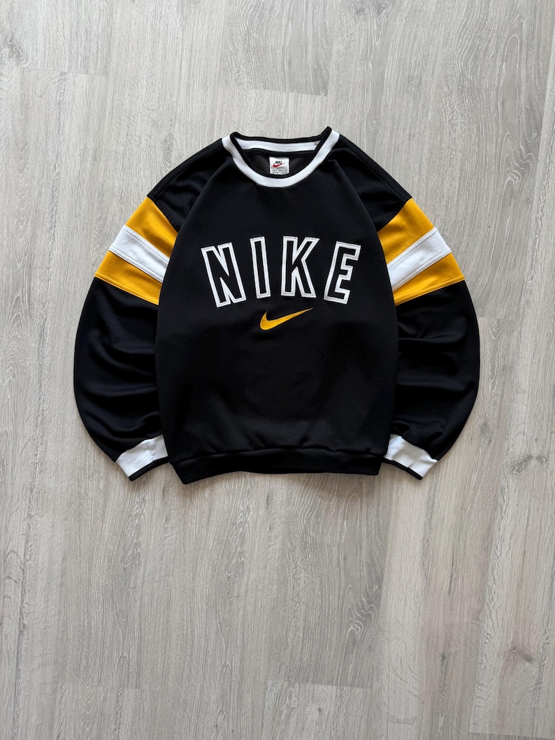Puede incluir: Sudadera negra Nike con ribetes blancos y rayas amarillas y blancas en las mangas. La palabra "NIKE" est&aacute; impresa en letras blancas grandes en el pecho, con un logotipo de Nike swoosh amarillo debajo.