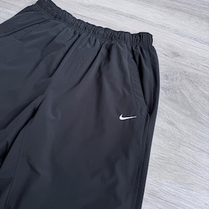 Puede incluir: Pantalones deportivos negros con cintura el&aacute;stica y bolsillos laterales. Un peque&ntilde;o logotipo blanco de Nike es visible en la pierna izquierda. Los pantalones parecen estar hechos de un material ligero y resistente al agua.