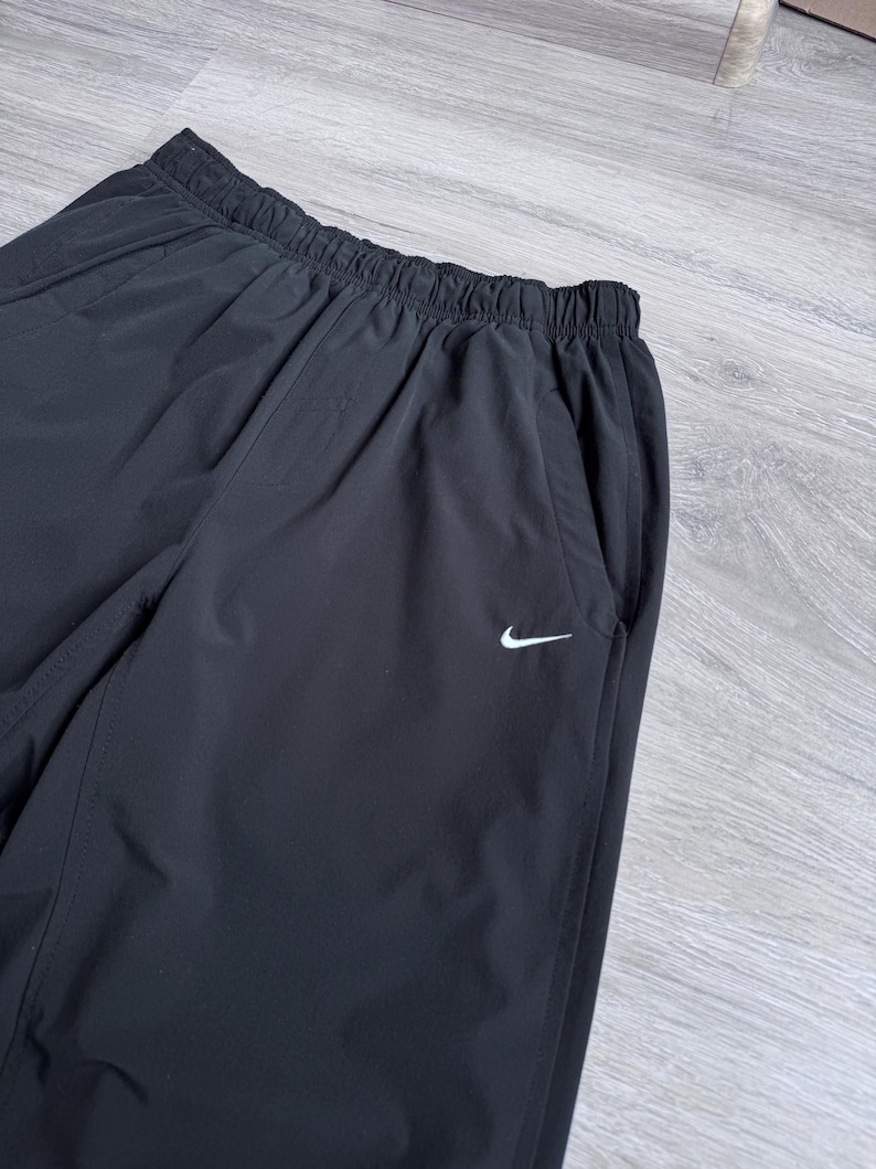 Puede incluir: Pantalones deportivos negros con cintura el&aacute;stica y bolsillos laterales. Un peque&ntilde;o logotipo blanco de Nike es visible en la pierna izquierda. Los pantalones parecen estar hechos de un material ligero y resistente al agua.