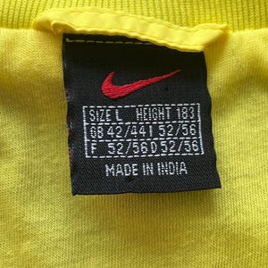 May include: Close-up of a yellow t-shirt tag with the Nike logo in red. The tag is black with white text indicating size, height, and measurements. The tag also states "MADE IN INDIA".