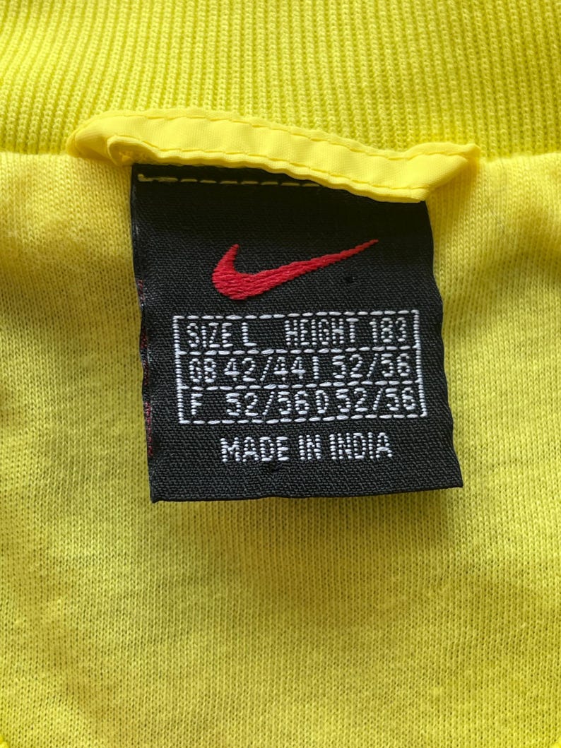May include: Close-up of a yellow t-shirt tag with the Nike logo in red. The tag is black with white text indicating size, height, and measurements. The tag also states "MADE IN INDIA".