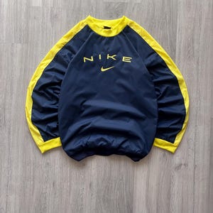 May include: Navy blue Nike pullover with yellow accents. The word "NIKE" is printed in yellow across the chest, with the Nike swoosh logo below. The collar and side panels are yellow.