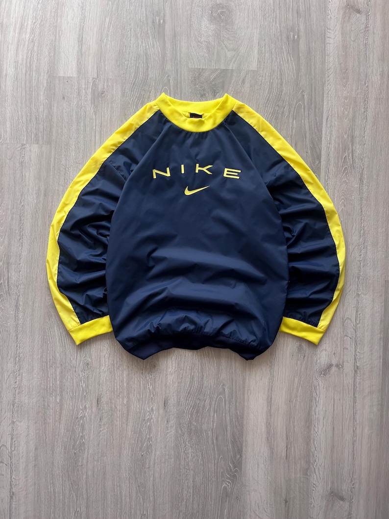 May include: Navy blue Nike pullover with yellow accents. The word "NIKE" is printed in yellow across the chest, with the Nike swoosh logo below. The collar and side panels are yellow.