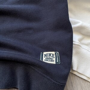 May include: Close-up of a navy blue sweatshirt with a ribbed hem and cuffs. A rectangular patch on the upper left chest reads "NIKE Athletic SINCE 1972". A portion of a light beige sweatshirt is visible on the right.