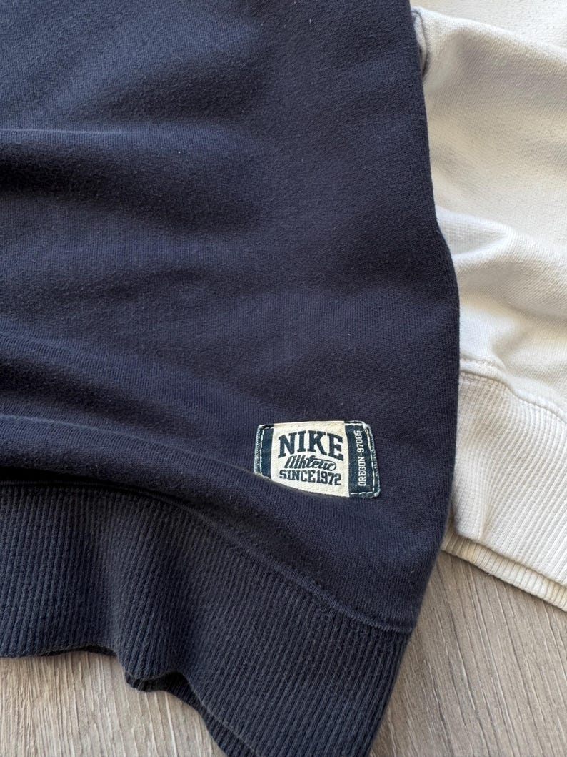 May include: Close-up of a navy blue sweatshirt with a ribbed hem and cuffs. A rectangular patch on the upper left chest reads "NIKE Athletic SINCE 1972". A portion of a light beige sweatshirt is visible on the right.