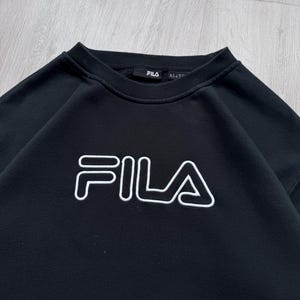 Peut inclure: Sweat-shirt noir &agrave; col rond avec le logo FILA blanc sur la poitrine. Le nom de la marque est soulign&eacute; en blanc. Le sweat-shirt a une encolure c&ocirc;tel&eacute;e et une &eacute;tiquette de marque FILA.