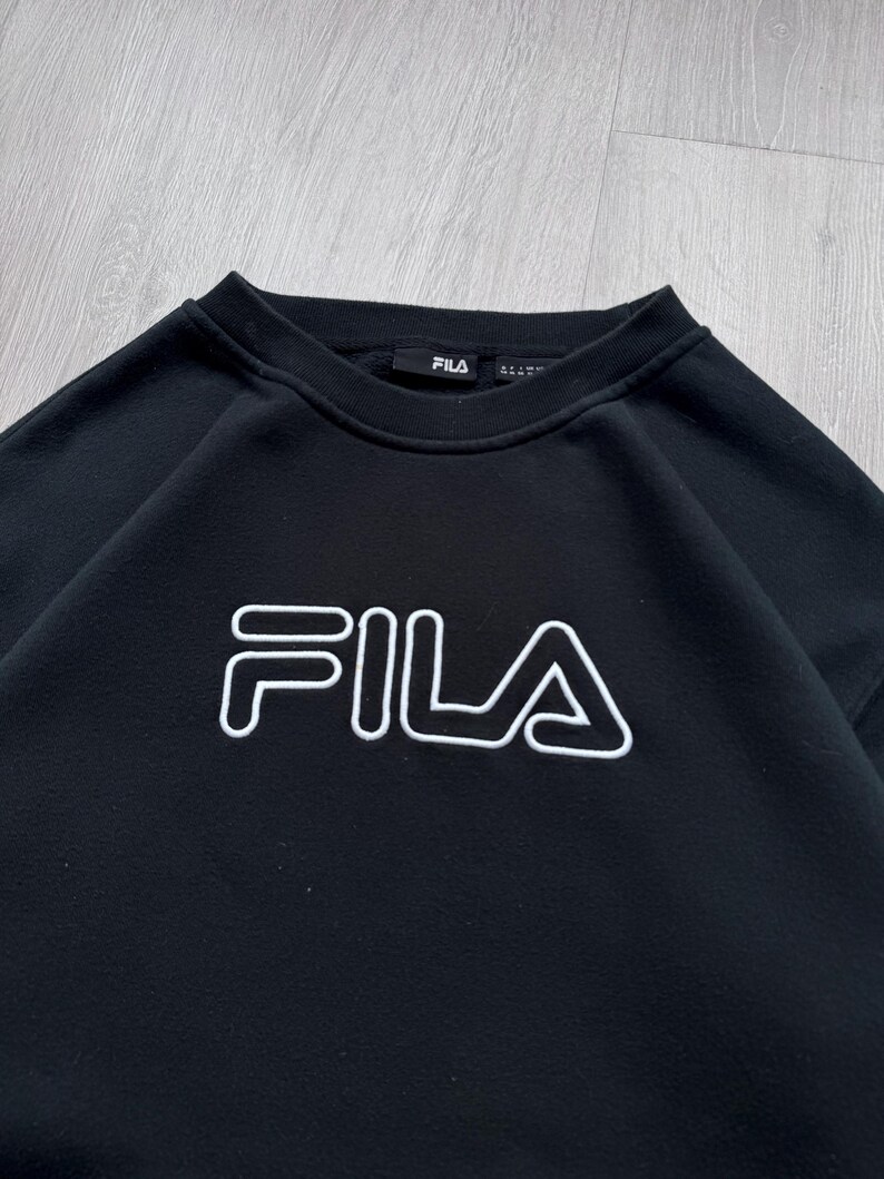 Peut inclure: Sweat-shirt noir &agrave; col rond avec le logo FILA blanc sur la poitrine. Le nom de la marque est soulign&eacute; en blanc. Le sweat-shirt a une encolure c&ocirc;tel&eacute;e et une &eacute;tiquette de marque FILA.