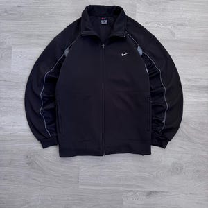 Puede incluir: Chaqueta deportiva negra con cremallera y logotipo blanco de Nike en el pecho. La chaqueta tiene cuello alto, bolsillos laterales y rayas grises en las mangas. Fabricada con un material atl&eacute;tico suave.