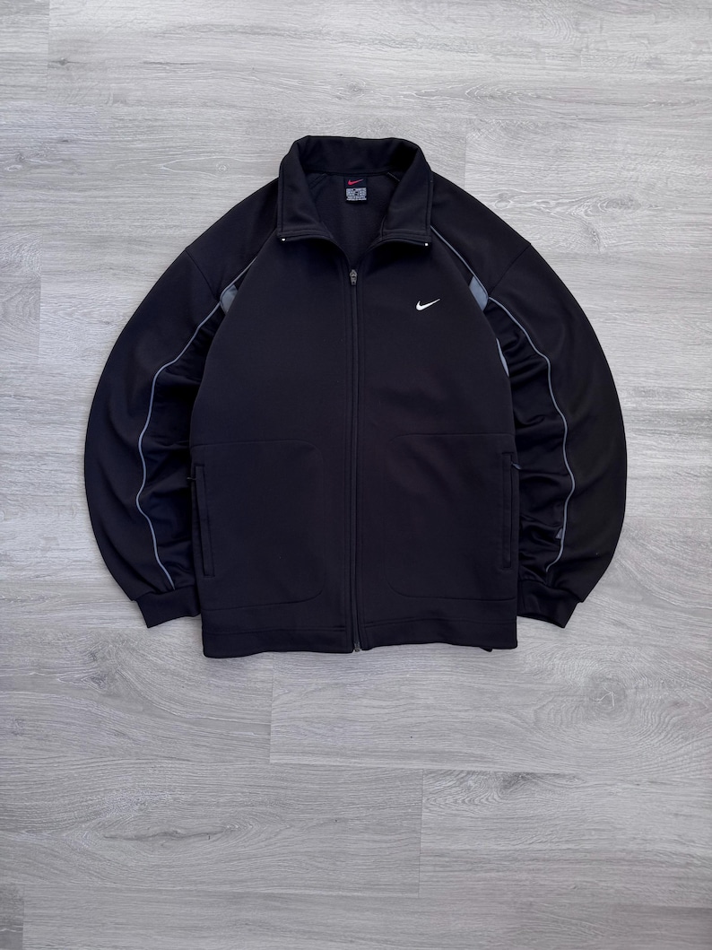 Puede incluir: Chaqueta deportiva negra con cremallera y logotipo blanco de Nike en el pecho. La chaqueta tiene cuello alto, bolsillos laterales y rayas grises en las mangas. Fabricada con un material atl&eacute;tico suave.