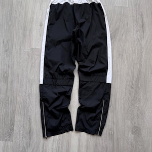 Peut inclure: Pantalon de sport noir avec des bandes lat&eacute;rales blanches et une taille blanche. Le pantalon a des fermetures &eacute;clair aux chevilles et un cordon de serrage. Le pantalon est en mati&egrave;re l&eacute;g&egrave;re.