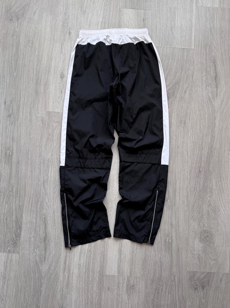 Peut inclure: Pantalon de sport noir avec des bandes lat&eacute;rales blanches et une taille blanche. Le pantalon a des fermetures &eacute;clair aux chevilles et un cordon de serrage. Le pantalon est en mati&egrave;re l&eacute;g&egrave;re.