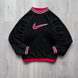 May include: Black sweatshirt with red and white striped detailing on the collar, cuffs, and hem. A red Nike swoosh logo, outlined in white, is on the chest. The long-sleeved top has a crew neck.